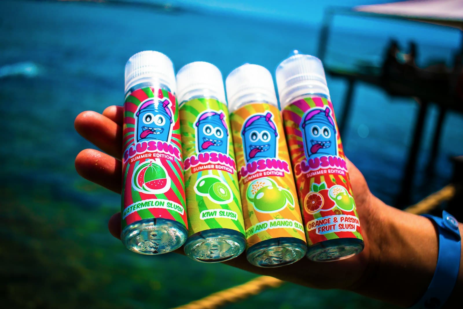 A hand holding four large capped e-liquid bottles with colourful fruit-flavour labels